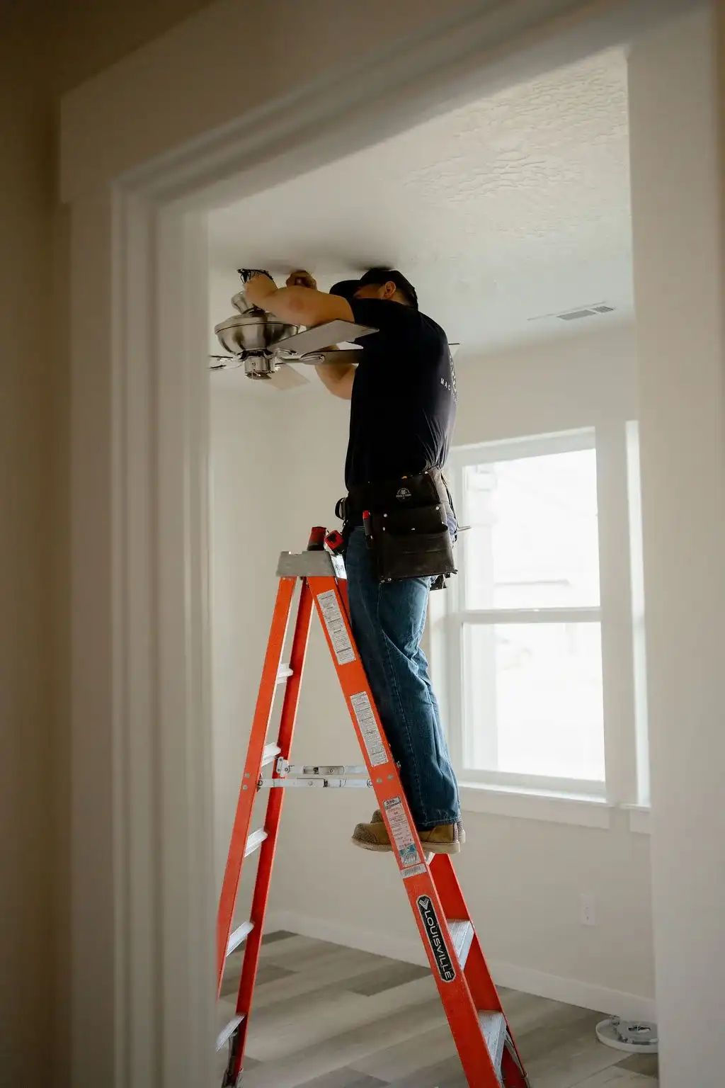 Electrician on an orange ladder installing a ceiling fan in a bright, empty room