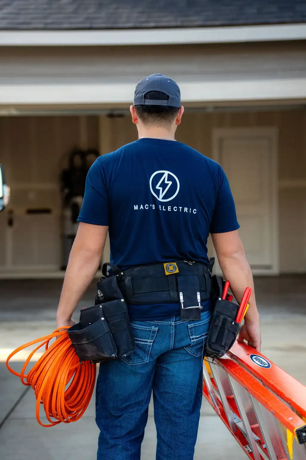 Electrician in a MAC's Electric shirt carrying an extension cord and a ladder into a garage